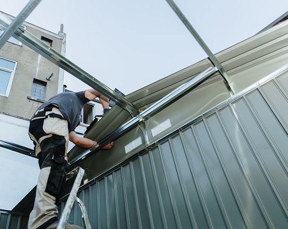 Installation toiture métallique : montage rapide de l'ossature. Montage d'une structure de toiture métallique. Un ouvrier sur une échelle fixe les panneaux de tôle ondulée sur l'ossature.