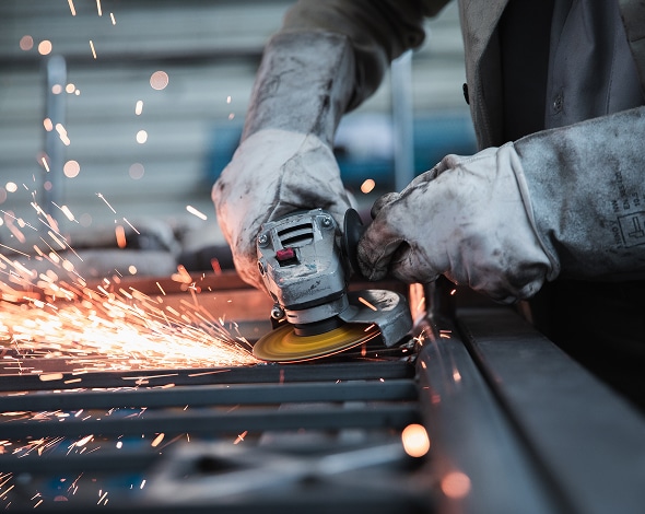 Meulage de métal en atelier : étincelles et sécurité. Gants de protection sur les mains d'un travailleur utilisant une meuleuse d'angle sur des profilés métalliques, créant un jet d'étincelles intense.